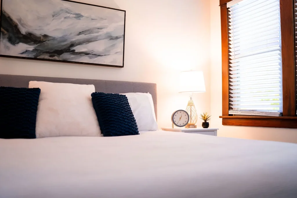 A bedroom with a bed, black and white pillows, a nightstand with a clock, a lamp, a small plant, a window with blinds, and abstract artwork on the wall.
