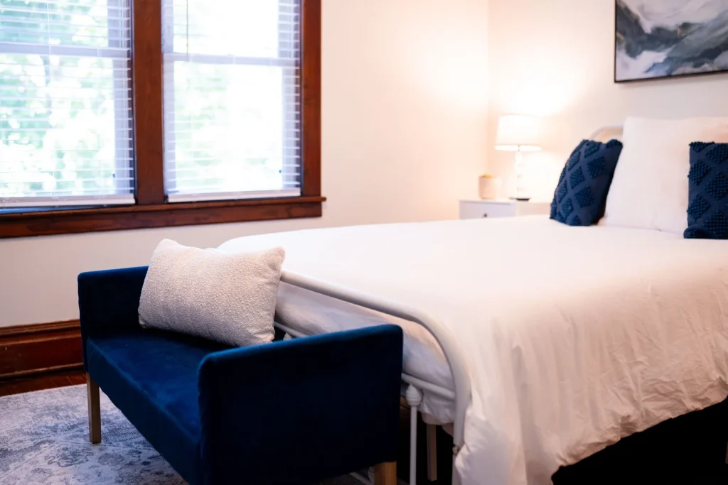 A bedroom with a large bed covered in white bedding, two navy blue pillows, a small bench at the foot of the bed, and a nightstand with a lamp.