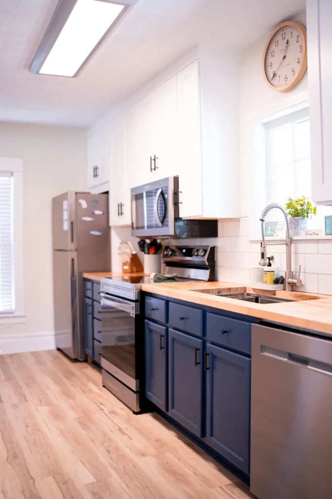 A modern kitchen with white upper cabinets, blue lower cabinets, a wooden countertop, stainless steel appliances, a window above the sink with a green plant, and a round wall clock.