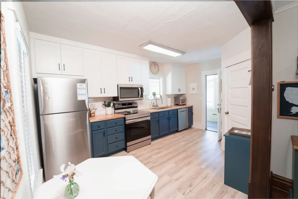 A bright kitchen with white upper cabinets, blue lower cabinets, stainless steel appliances, a window above the sink, and a wall clock.