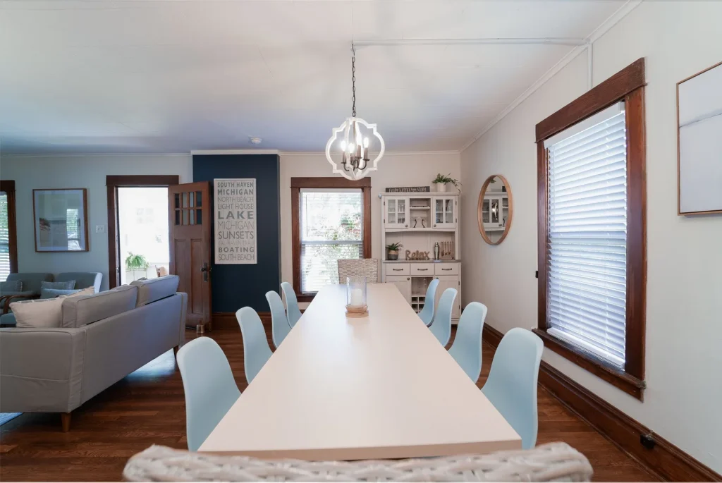 Dining area with a white table and chairs, a white china cabinet, and large windows with wooden trim, in a coastal-themed home.