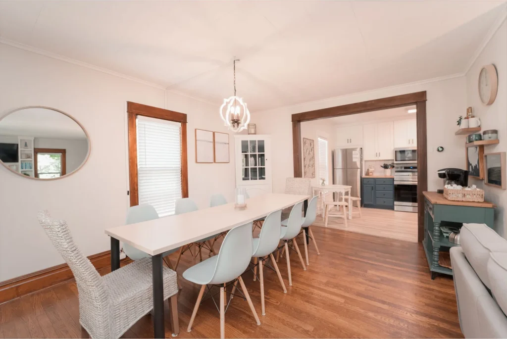 Dining area with a long white table, white and wicker chairs, hardwood floors, and a large mirror on the wall, opening to a kitchen with white cabinets, a stainless steel refrigerator, and gas stove.