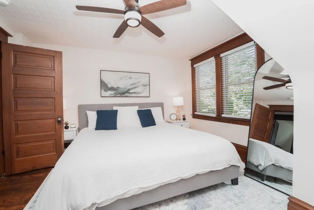 A bedroom with a white bed, gray headboard, and navy pillows. Brown wooden door and window trim, with blinds. A nightstand with a lamp, clock, and small plant. A ceiling fan and an arched mirror reflecting part of the room.