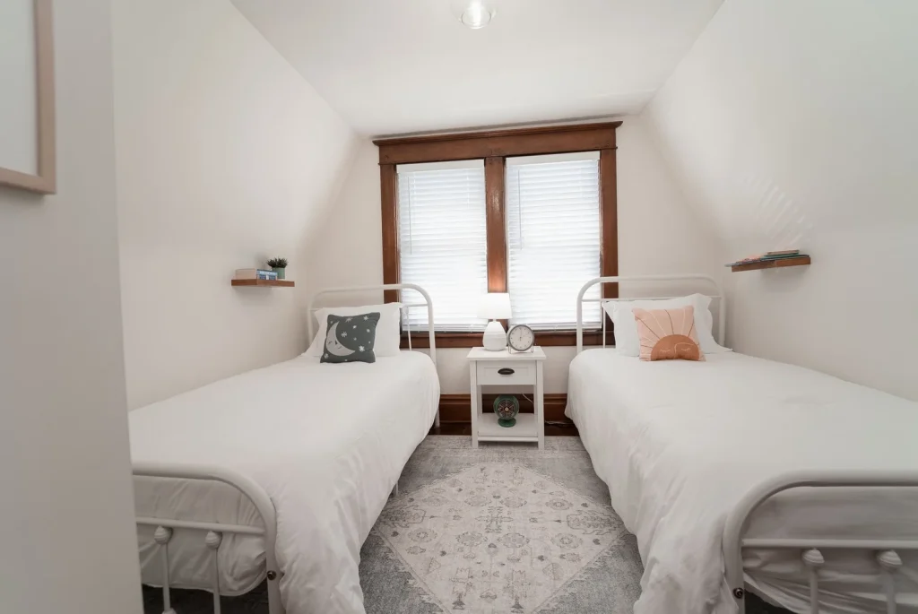 A bedroom with two single beds with white bedding, a nightstand in between, and large windows with wooden trim behind the beds.