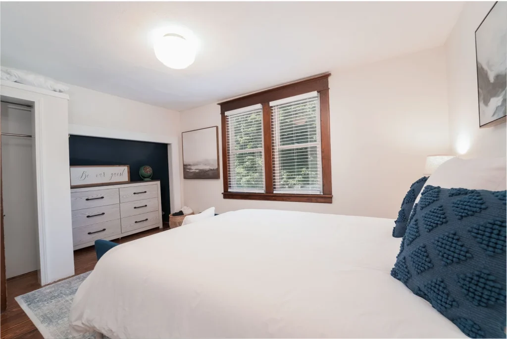 A bedroom with a large bed, blue textured pillows, wooden window frames, and a dresser with a sign that reads 'Be our guest.'